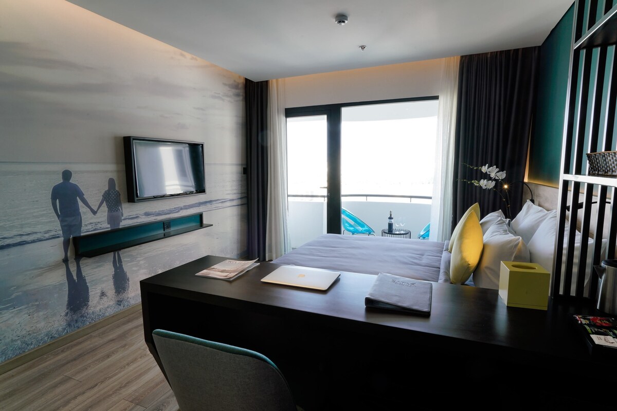 Quarto de hotel com uma cama, uma escrivaninha com um laptop, uma televisão e vista para o mar.