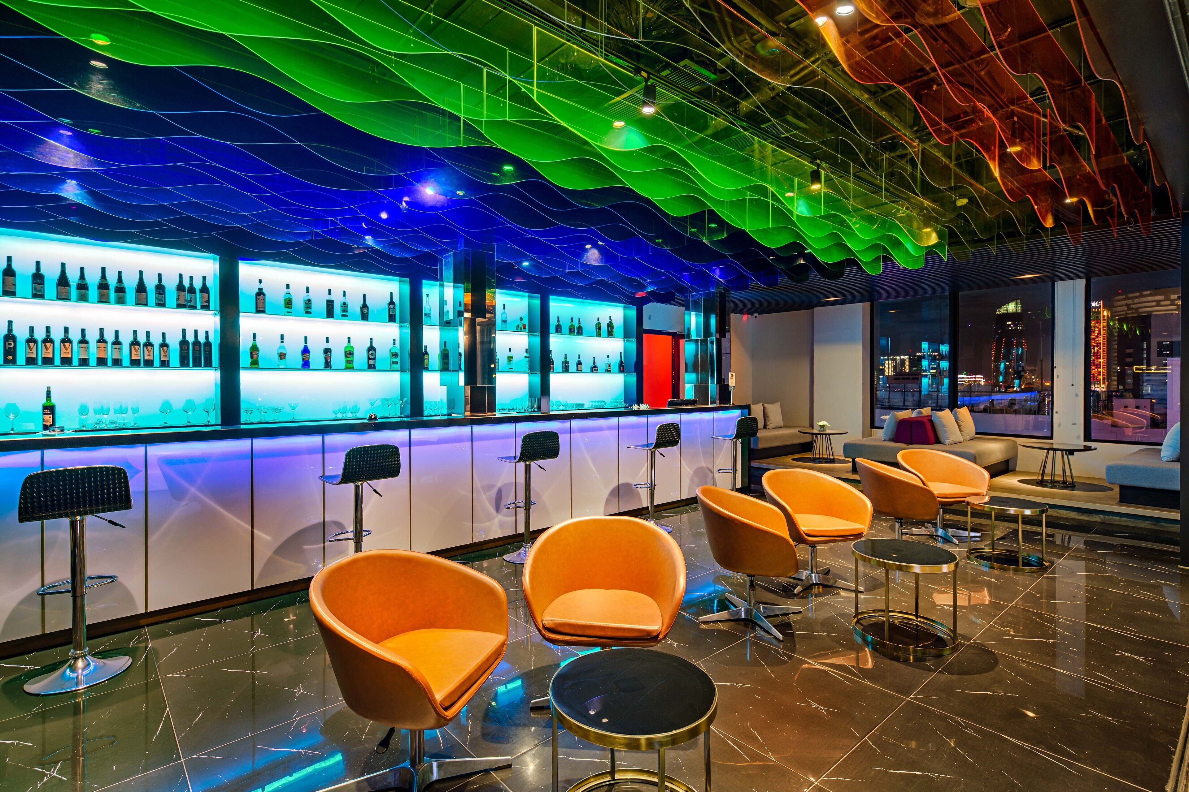 A modern bar area with a neon-lit ceiling, bar stools, and a seating area with orange chairs.