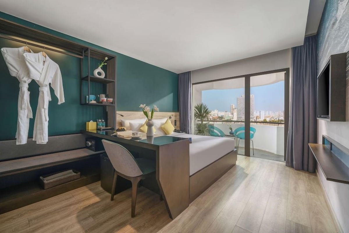 Quarto de hotel moderno com cama, escrivaninha, cadeira, televisão e varanda com vista para a cidade.