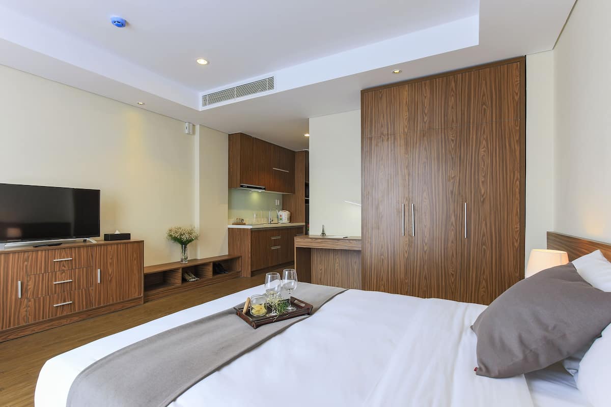 A modern hotel room with a large bed, wooden wardrobe, a flat-screen TV, a small kitchenette, and a bedside table with a lamp.