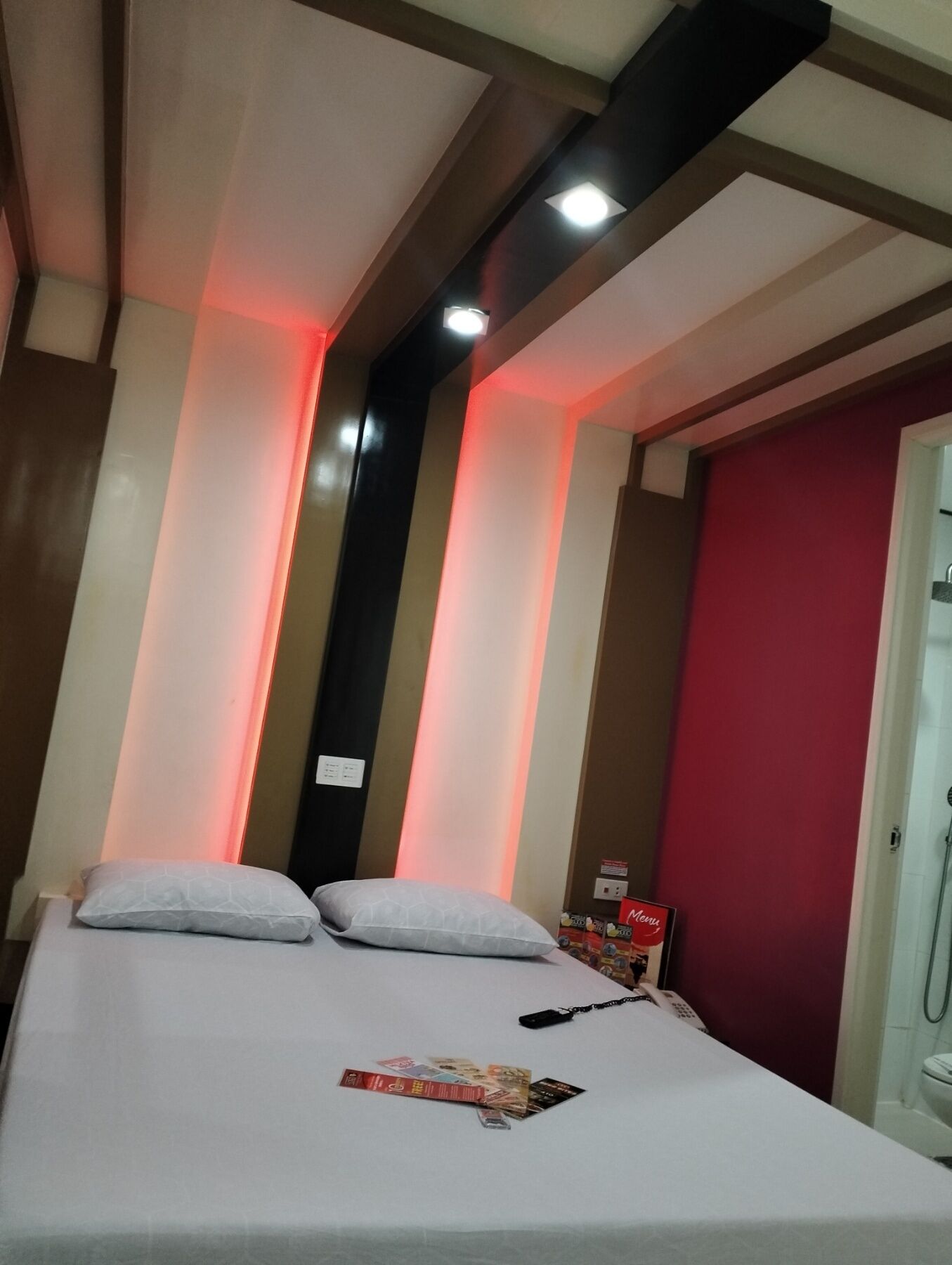 A hotel room with a bed, pillows, a nightstand, a red wall, and a shower area.