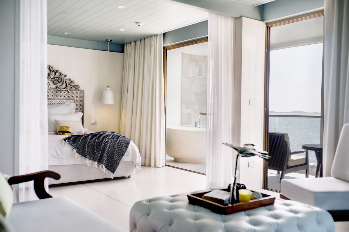 A modern hotel room with a bed, a sitting area with a chair and ottoman, a desk with a lamp, and a view of the sea through a large window.