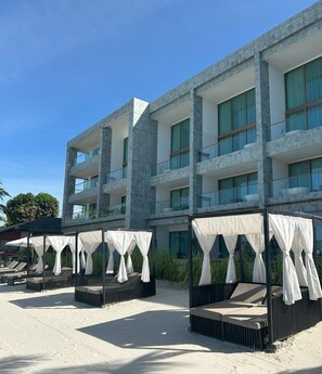 A modern hotel with multiple balconies, outdoor seating areas with white canopies, and a sandy beach in front.