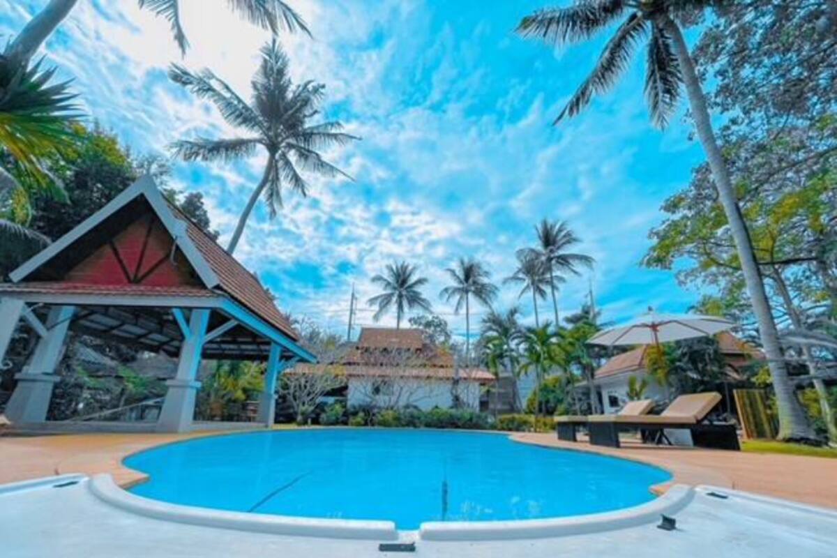 A swimming pool with a surrounding deck, palm trees, and a building with a red roof.