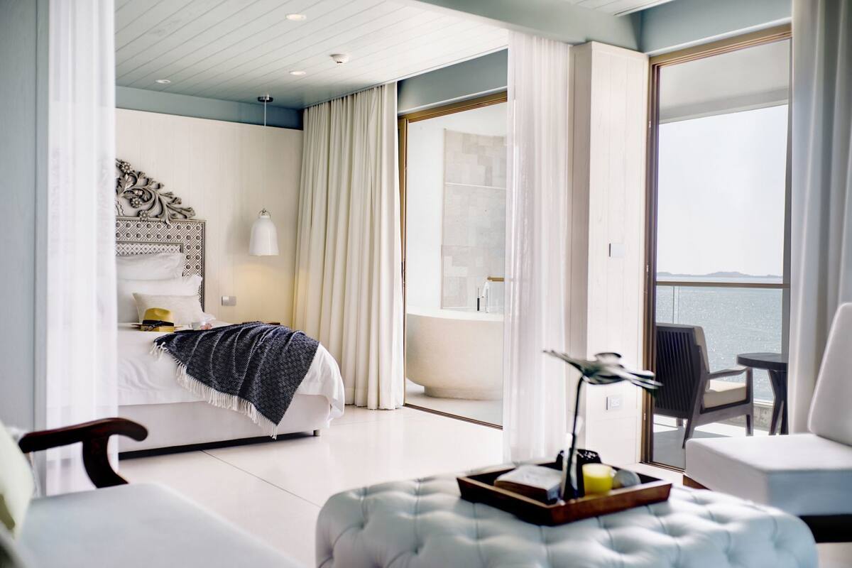 A modern hotel room with a bed, a seating area with a table and chairs, and a balcony with a view.