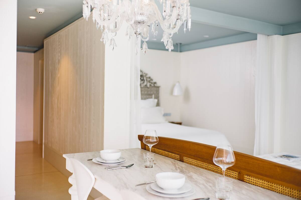 A hotel room with a dining table set for two, a bed with a wooden headboard, and a chandelier.