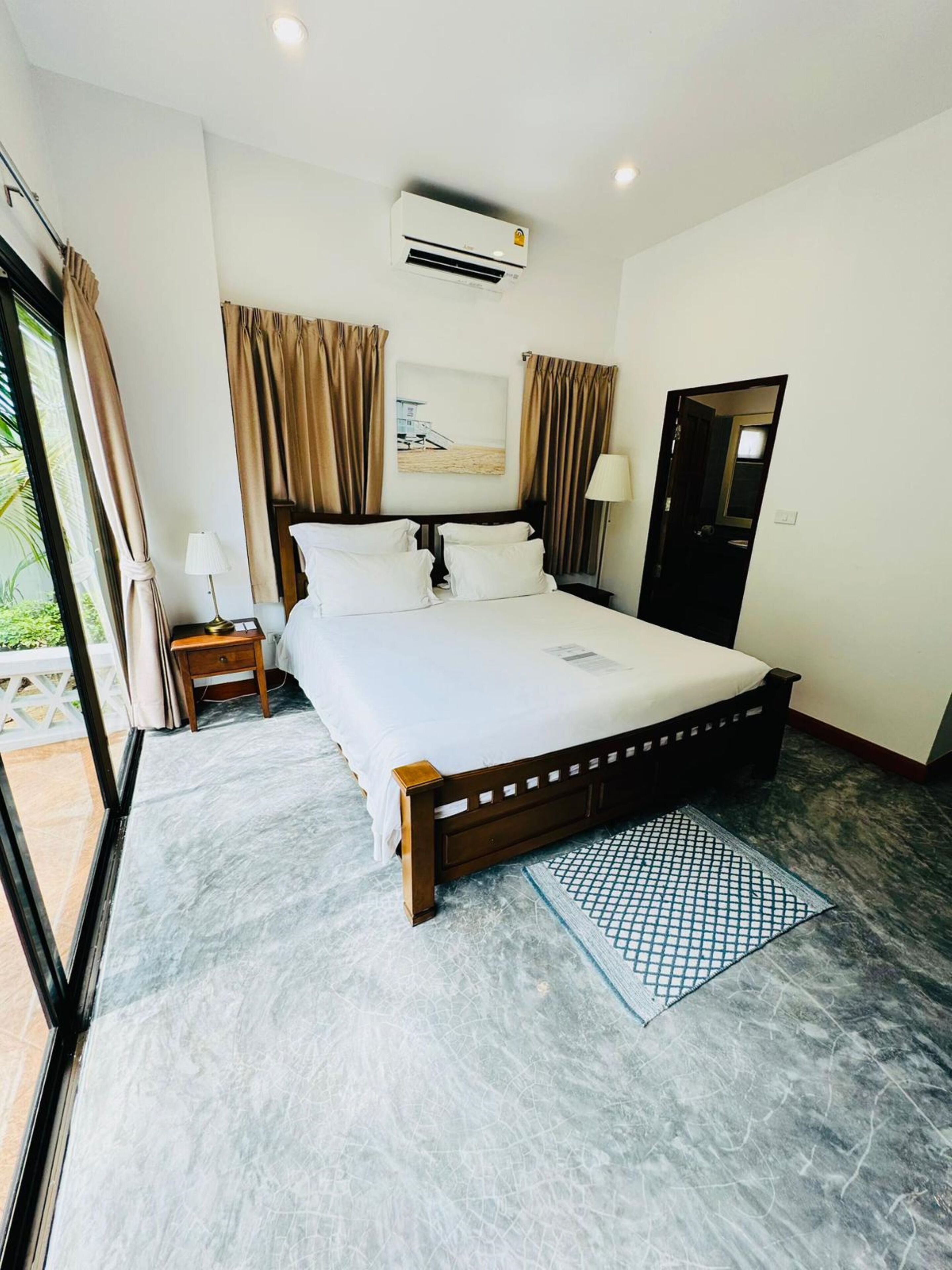 A hotel room with a large bed, a nightstand, a lamp, an air conditioning unit, and a view of the outdoors.