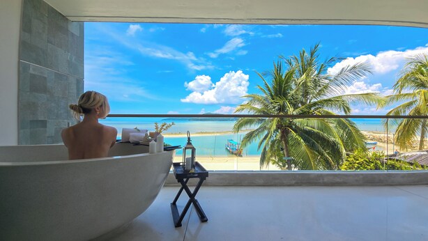 A person relaxing in a large bathtub with a view of a beach and palm trees. - The Beach Samui (Koh Samui)