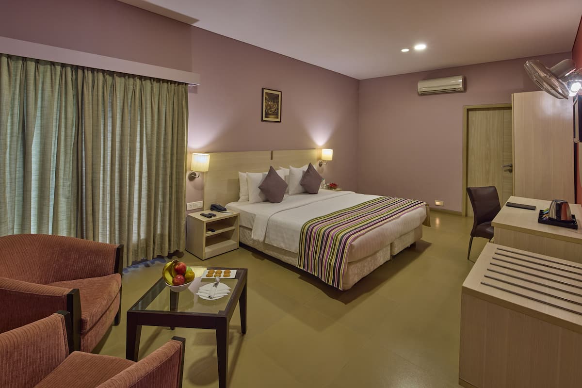A hotel room with a bed, a desk, a chair, a table with fruit, and a window with curtains.