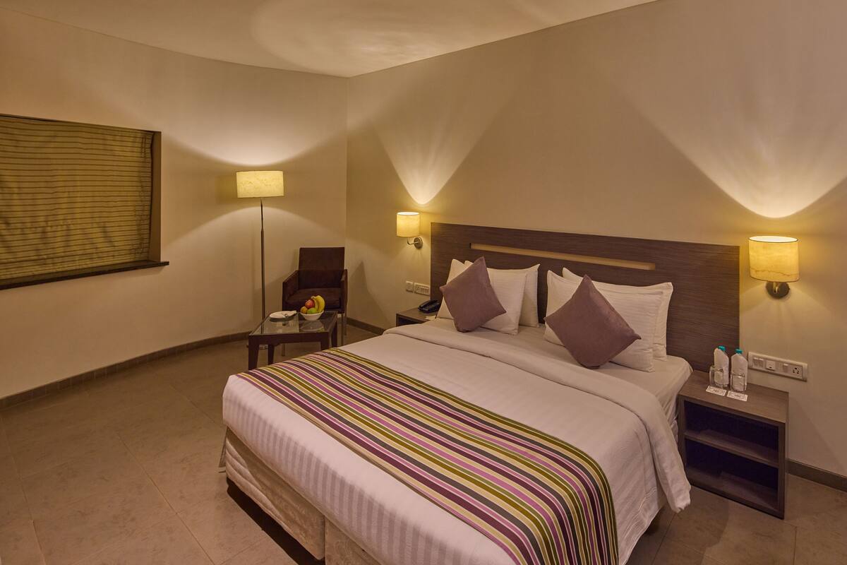 A hotel room with a large bed, two bedside tables, a chair, a bedside lamp, and a window with blinds.
