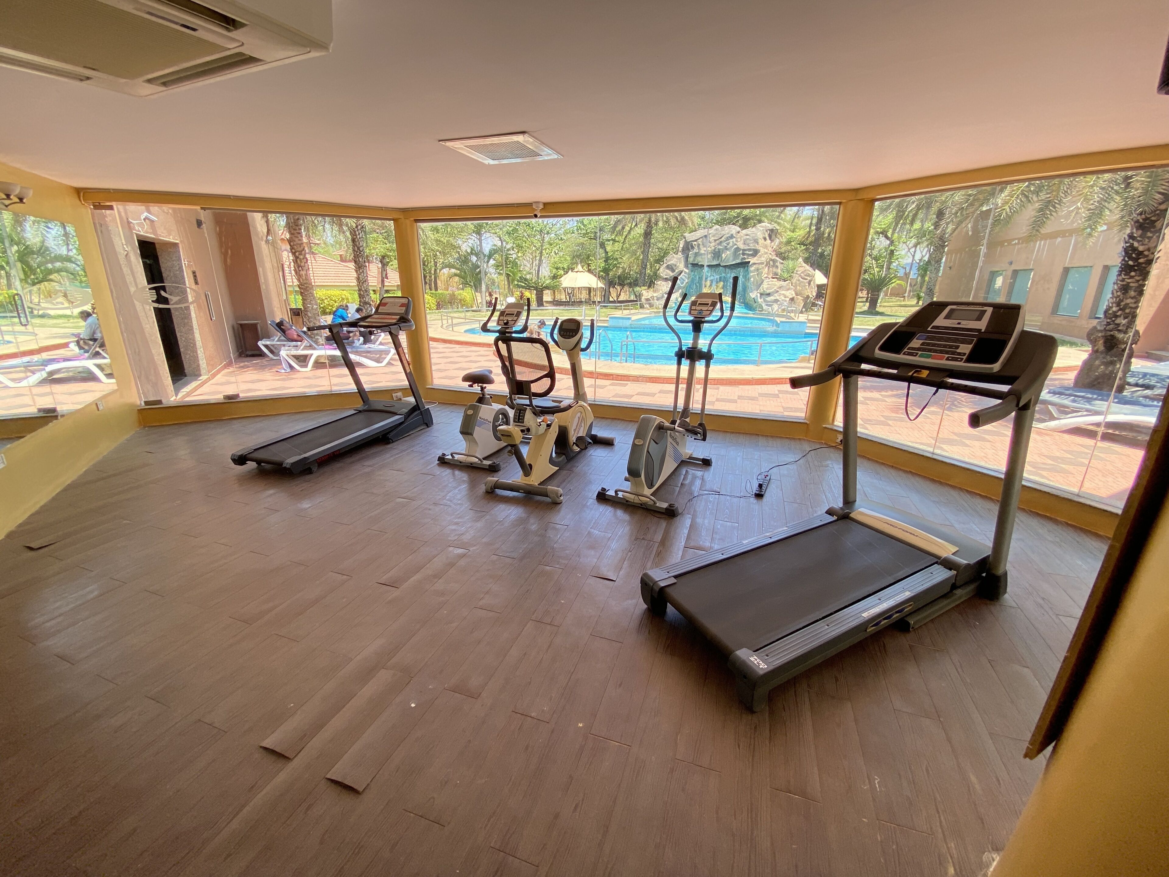 A gym with treadmills, an elliptical, and a stationary bike, with large windows overlooking a pool and outdoor area.