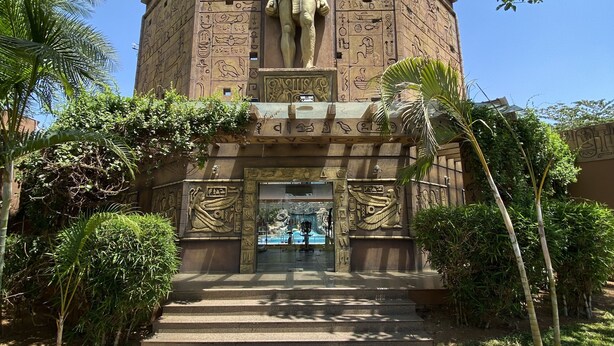 A large, ancient Egyptian-style building with a prominent statue at the entrance, surrounded by greenery. - Golden Amoon Resort (Hoskote)