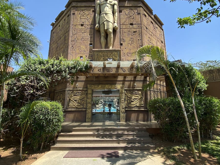 A large, ancient Egyptian-style building with a prominent statue at the entrance, surrounded by greenery.