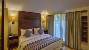 A hotel room with a large bed, wooden headboard, two bedside tables with lamps, a nightstand, and a large window with curtains. - Golden Amoon Resort (Hoskote)