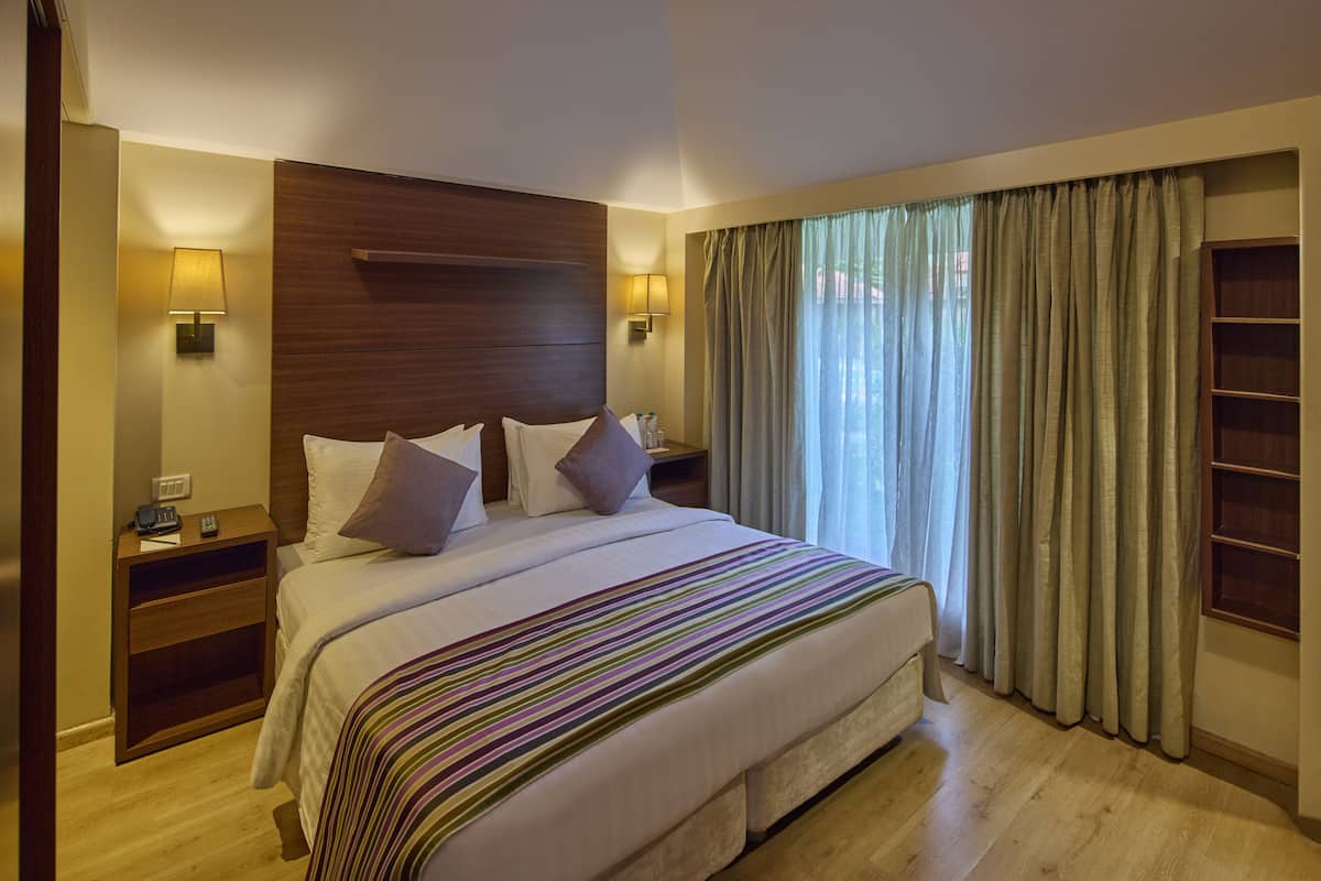 A hotel room with a large bed, wooden headboard, two bedside tables with lamps, a nightstand, and a large window with curtains.