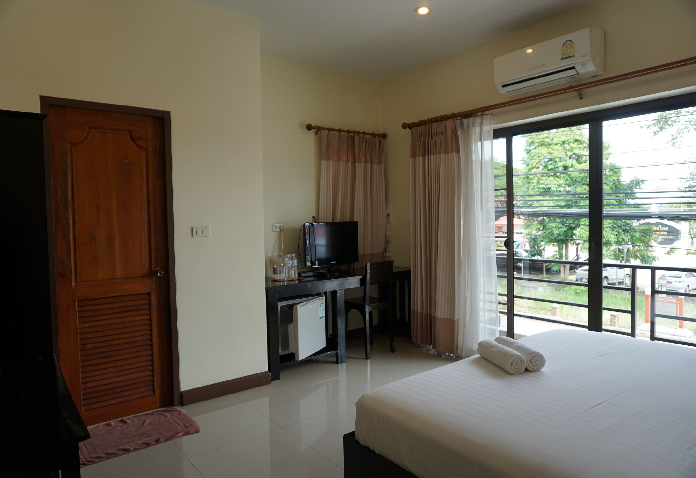 A hotel room with a bed, a desk with a television, a window with curtains, and an air conditioning unit.