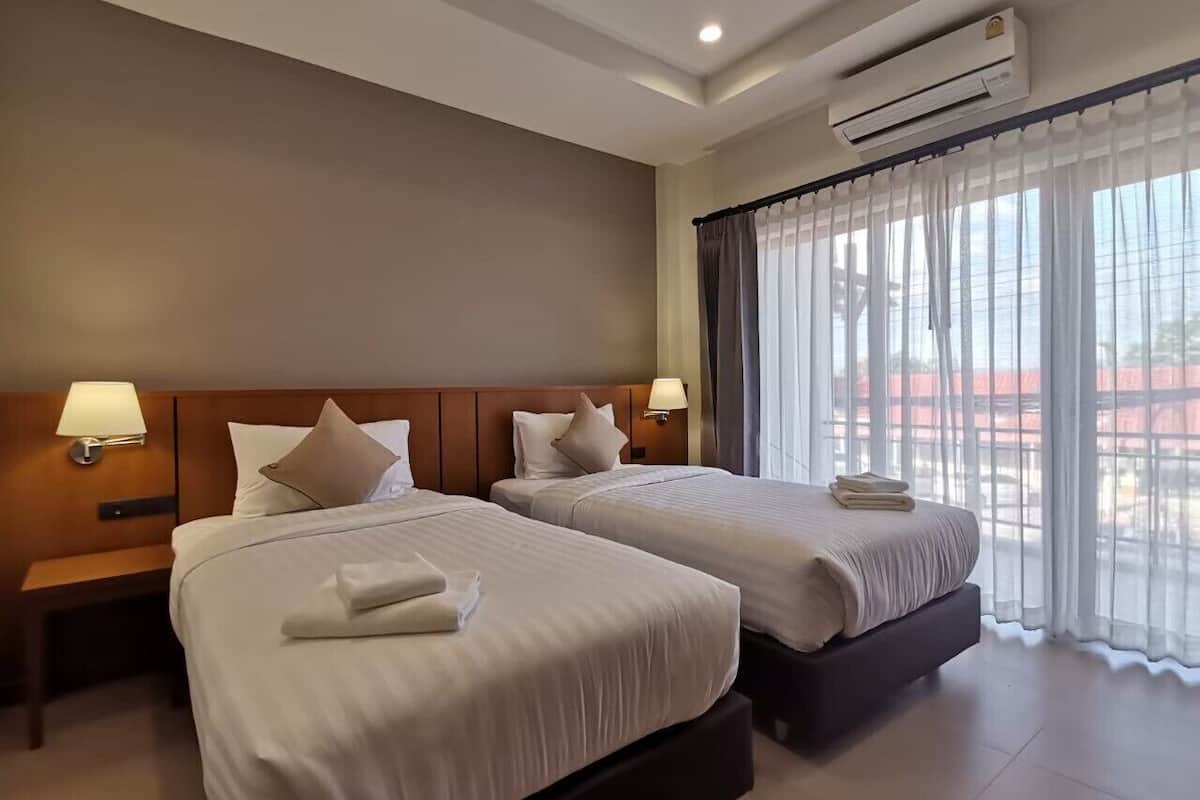 A hotel room with two beds, a nightstand, a lamp, an air conditioner, and a window with curtains.