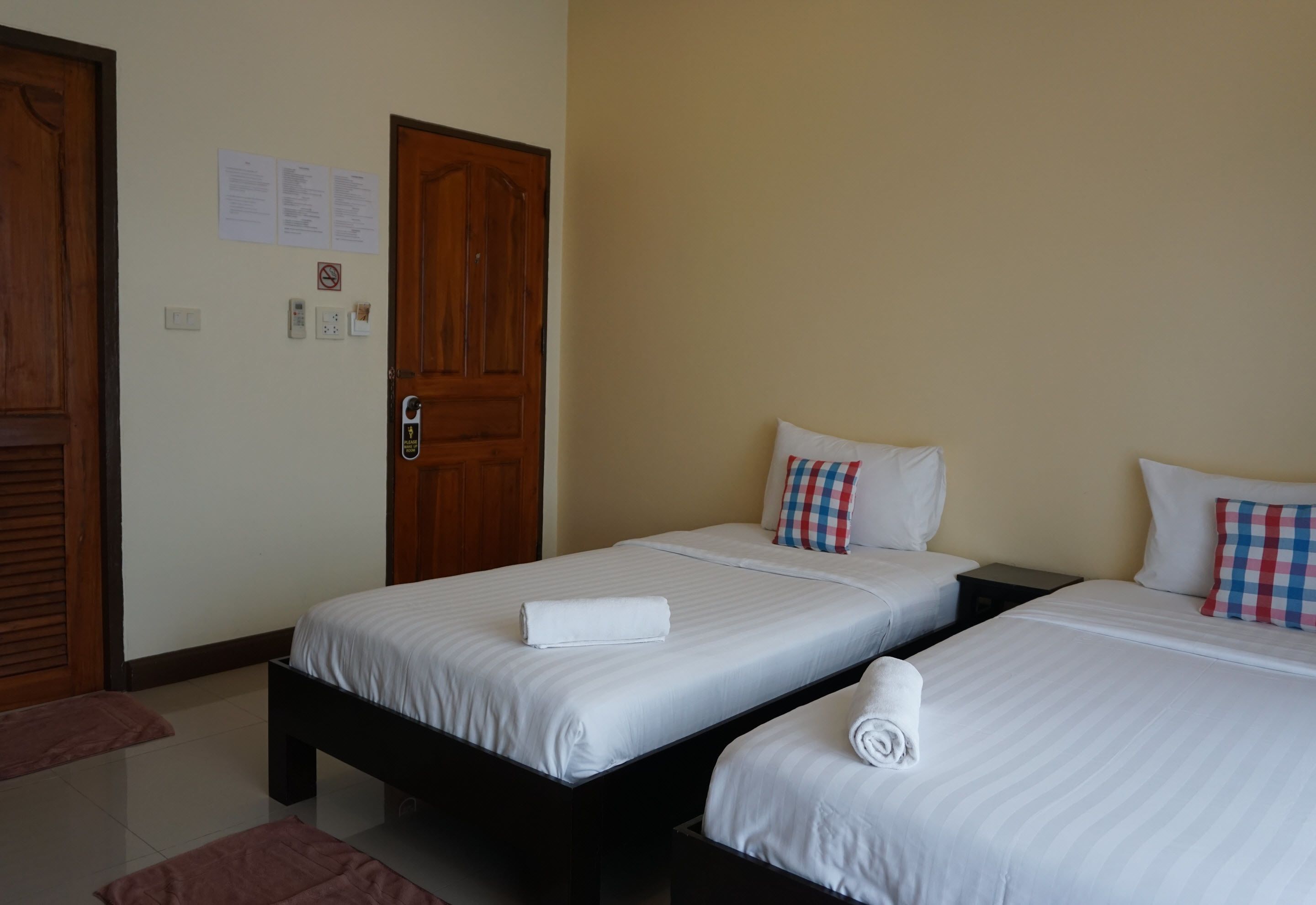 A hotel room with two single beds, wooden doors, a small table, and a wall-mounted light switch.