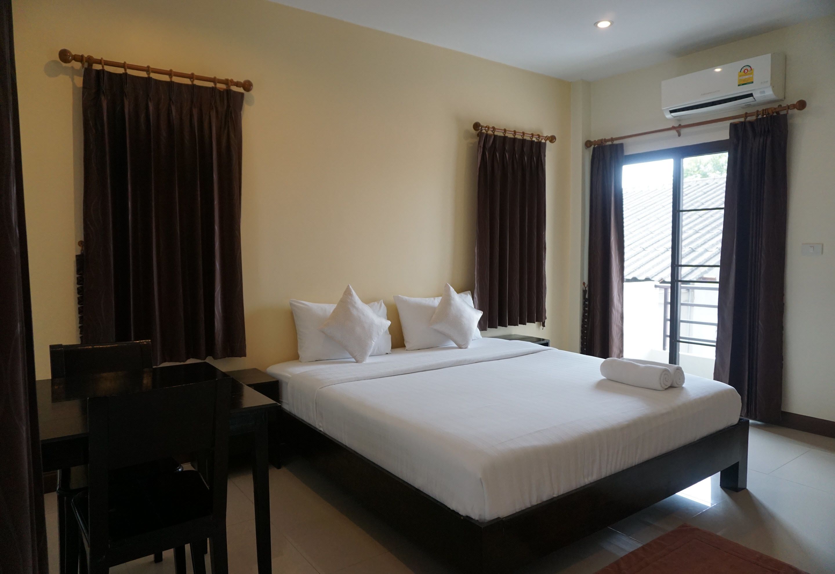 A hotel room with a large bed, two windows with curtains, a desk with a chair, and an air conditioning unit.