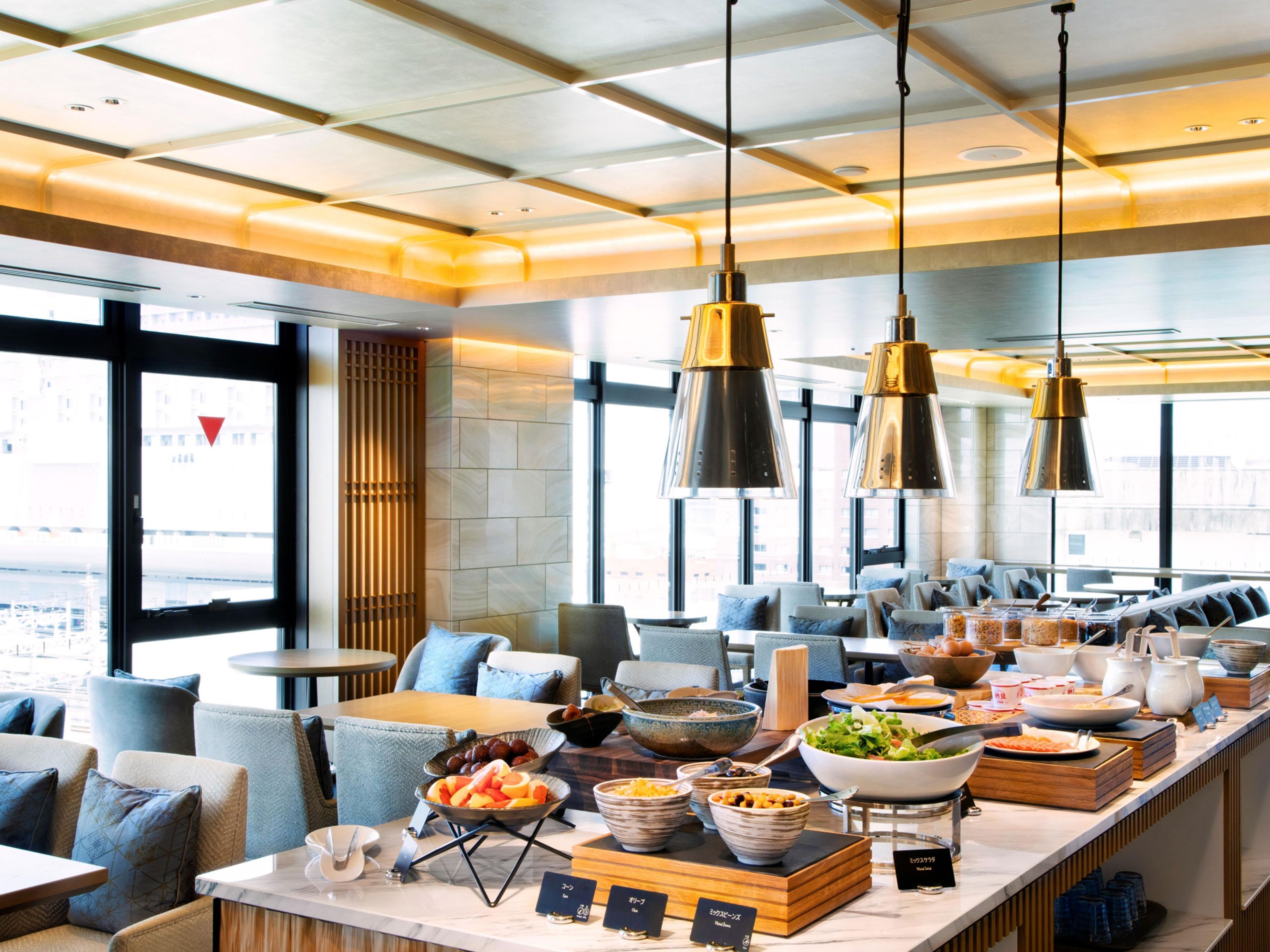 A modern hotel dining area with a buffet setup, seating arrangements, and large windows offering a view.