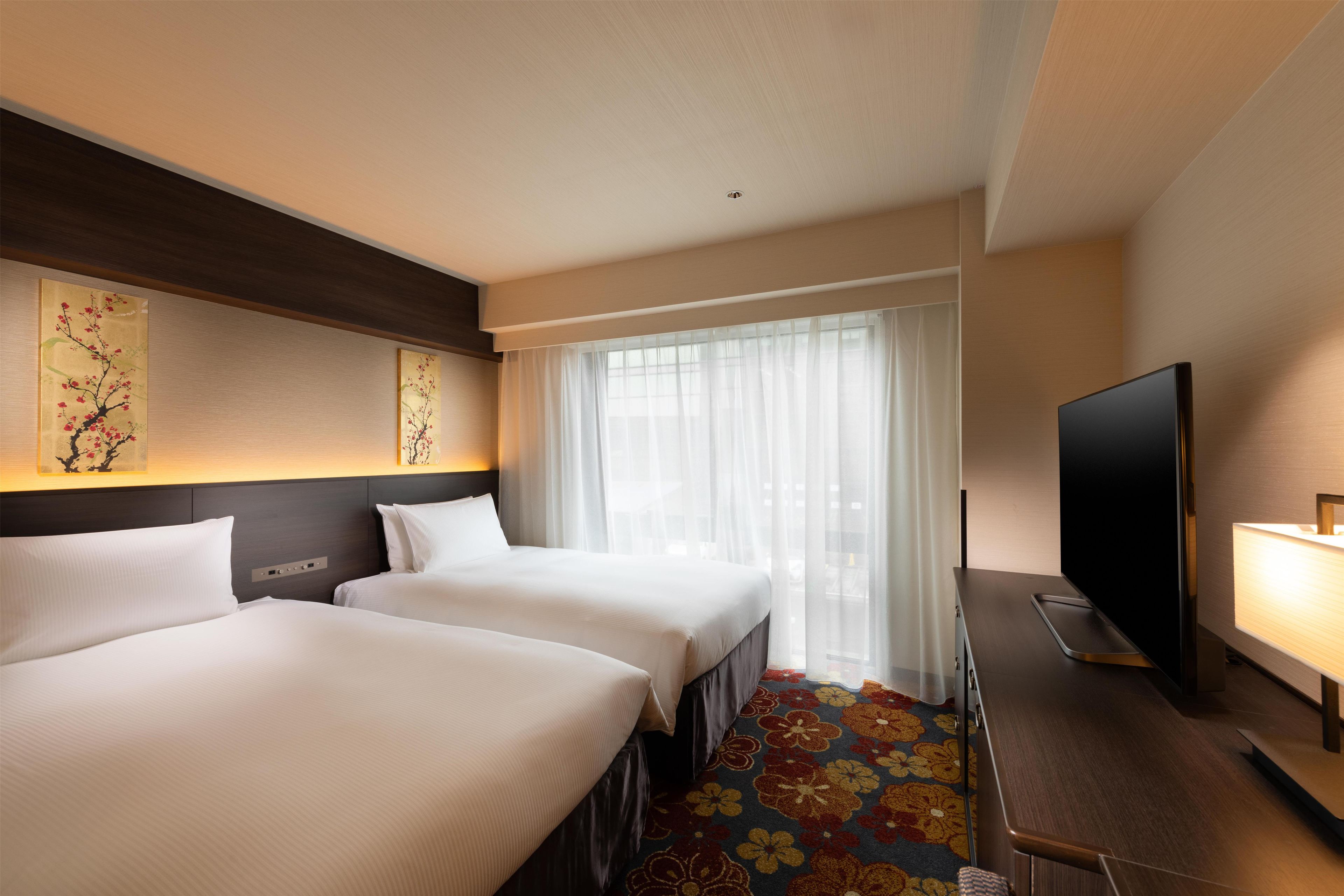 A hotel room with two beds, a television, a desk with a lamp, and a window with sheer curtains.