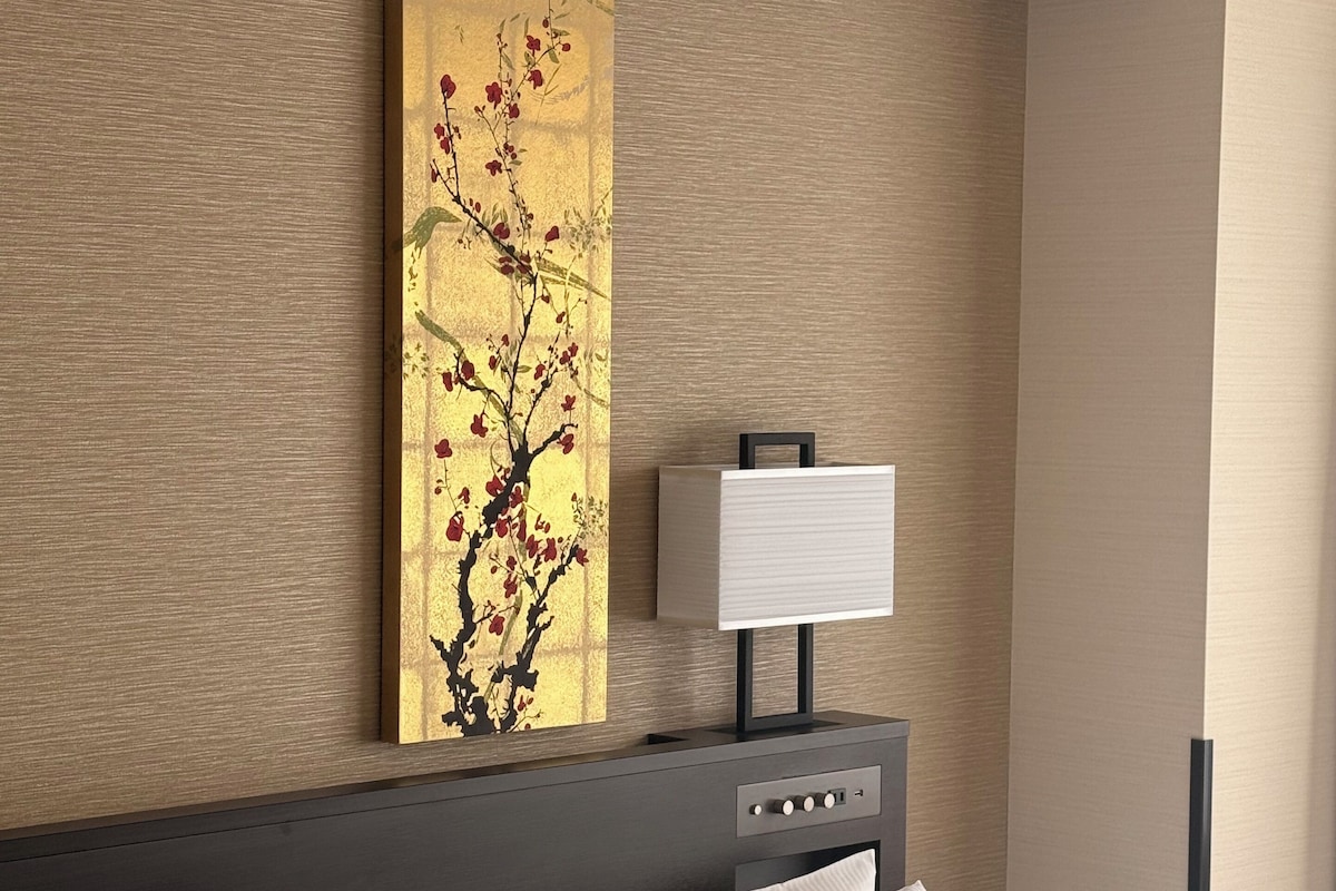 Camera d'albergo moderna con un ampio letto, un comodino con una lampada e un'opera d'arte a parete raffigurante un albero con fiori rossi.