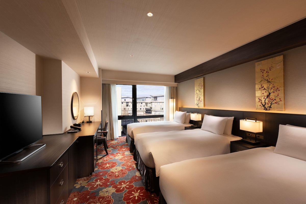 Habitación de hotel con dos camas, televisión, escritorio y vistas a edificios.