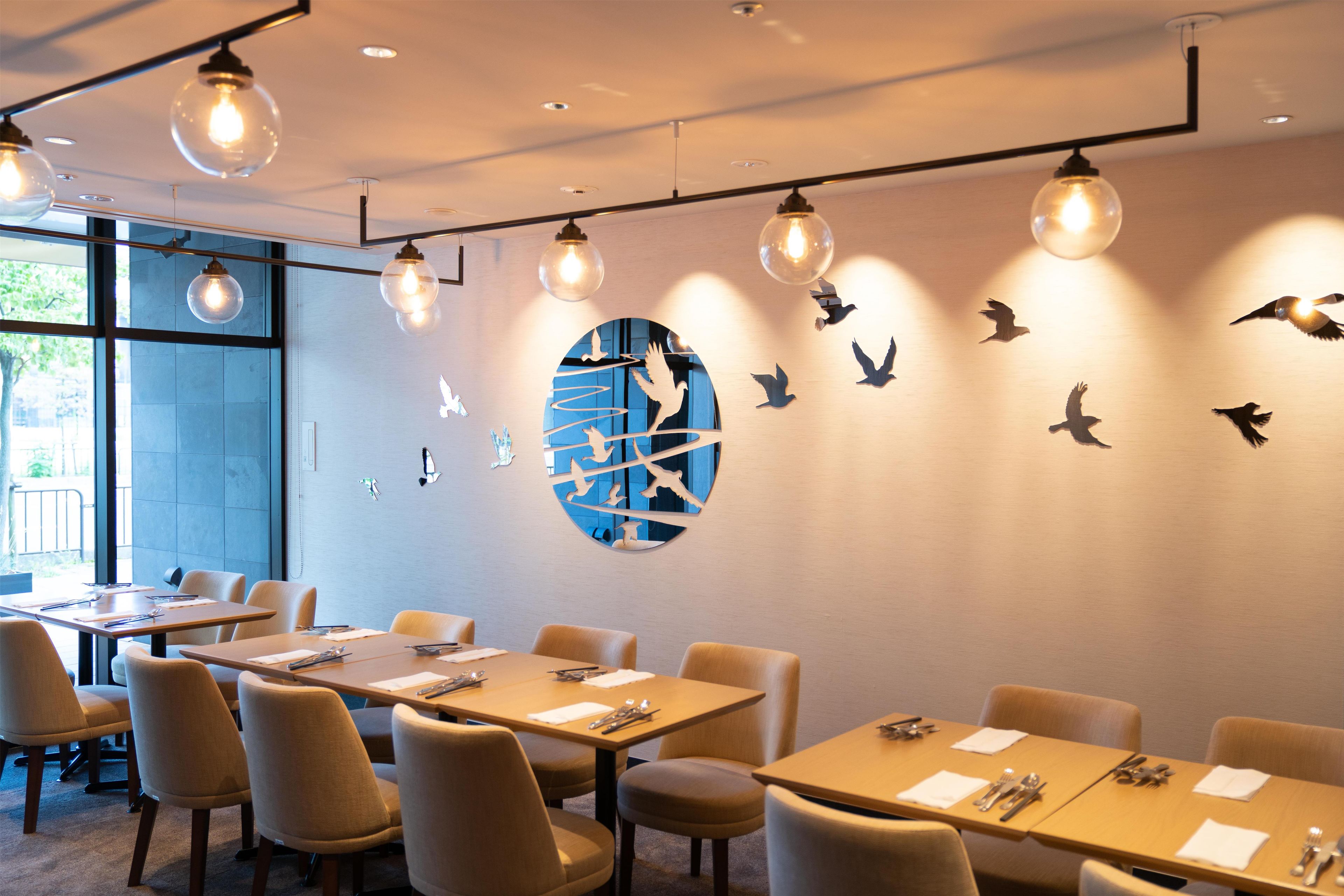 Un espace repas moderne avec des tables et des chaises, un mur décoré de motifs d’oiseaux découpés et des suspensions lumineuses.