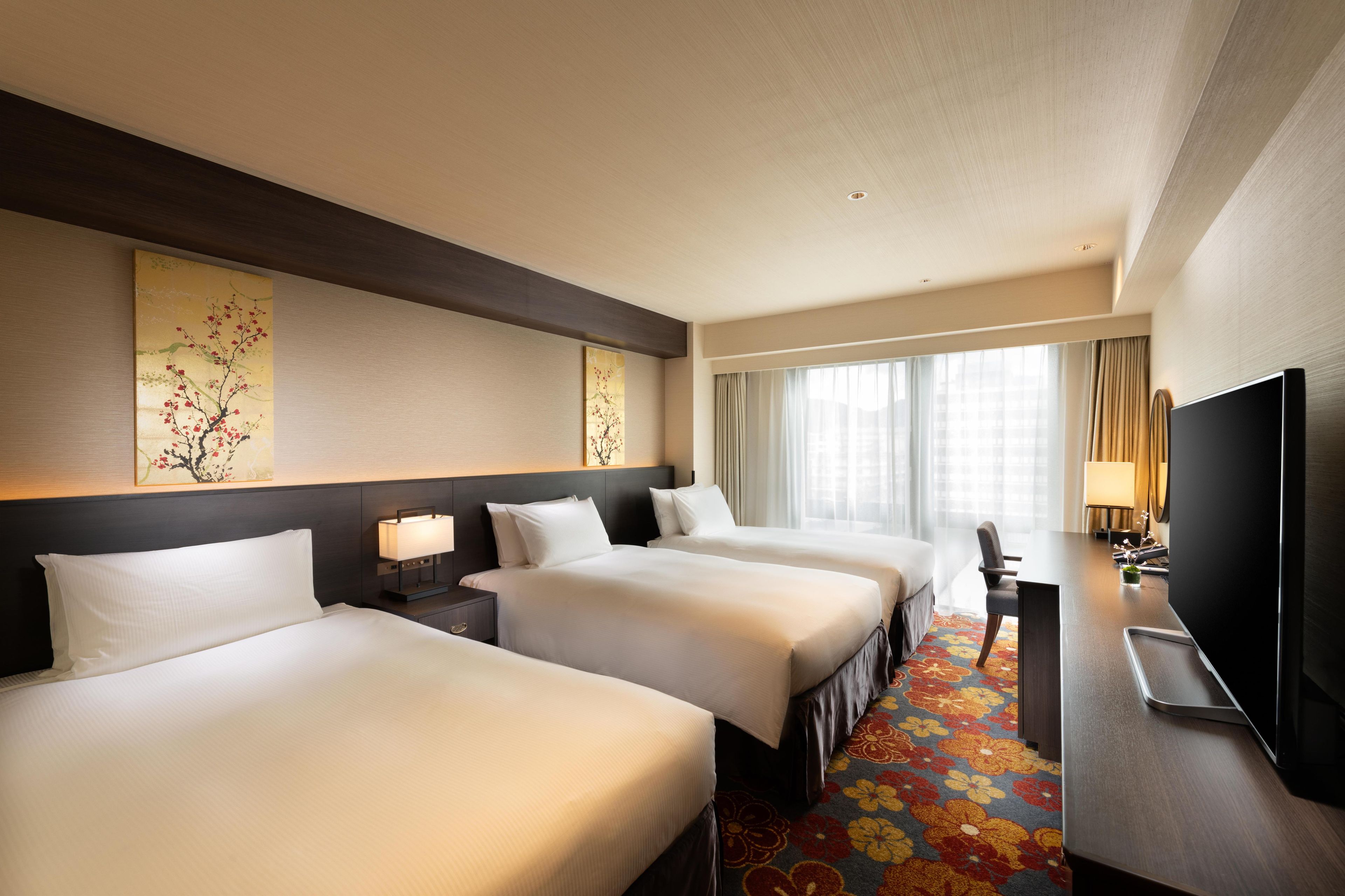 A hotel room with three beds, a desk, a TV, and artwork on the walls.