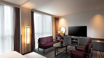 A hotel room with a bed, a sofa, a TV, a desk with a lamp, and a large window with curtains.