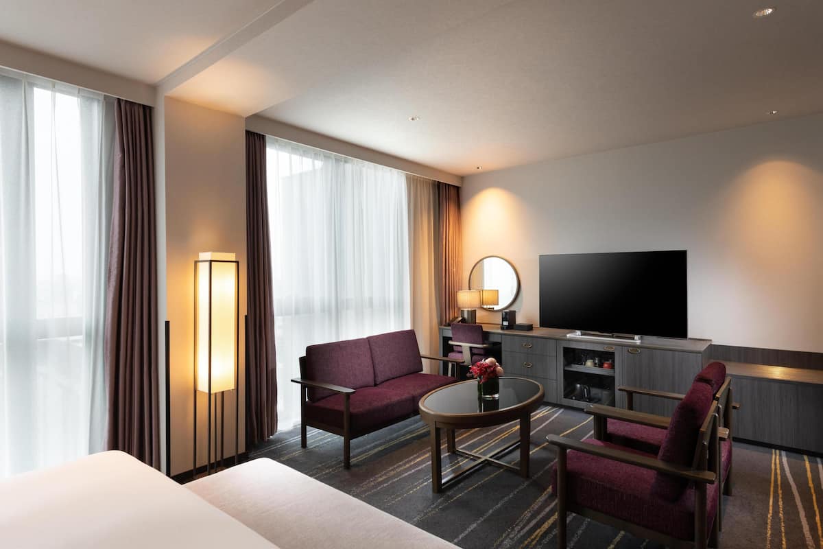 Una camera d'albergo con un letto, un divano, una televisione, una scrivania con una lampada e un'ampia finestra con tende.