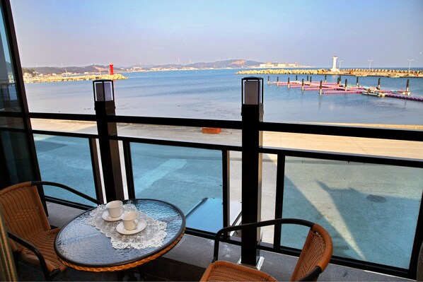 A balcony with wicker chairs and a table with cups, overlooking a calm sea and a pier with a lighthouse.