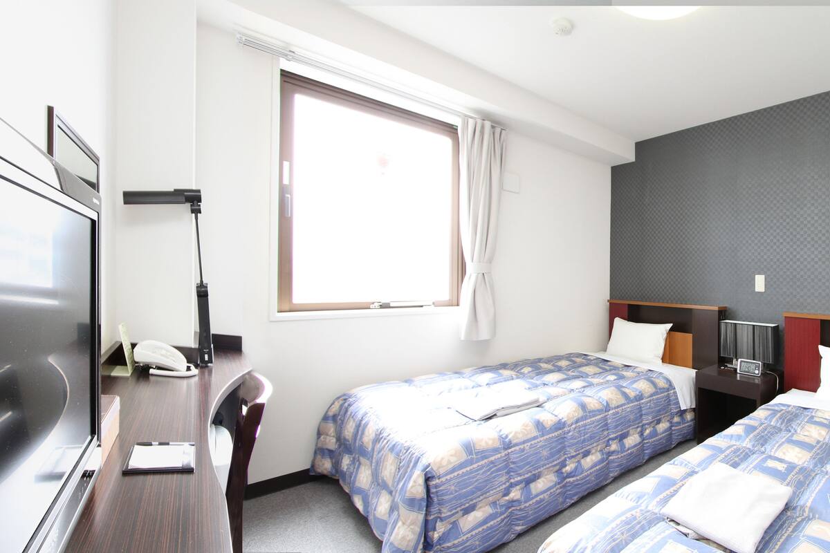 A hotel room with two beds, a desk, a television, and a window with curtains.