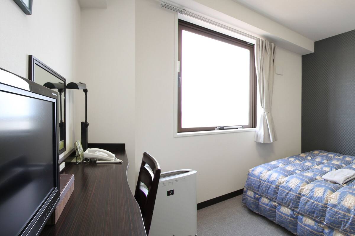 A hotel room with a bed, a desk with a telephone, a chair, a window with curtains, and a wall-mounted air conditioning unit.