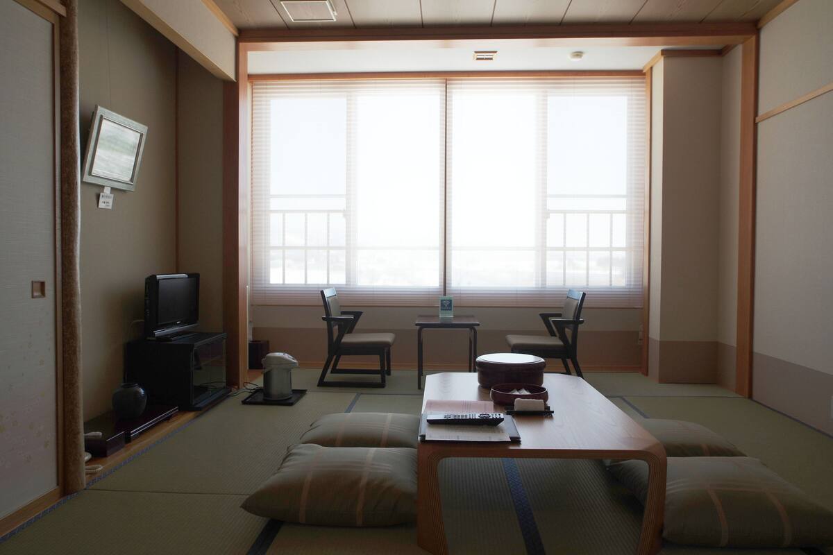 A traditional Japanese-style room with tatami flooring, a low table, cushions, a television, and a large window with sliding doors.