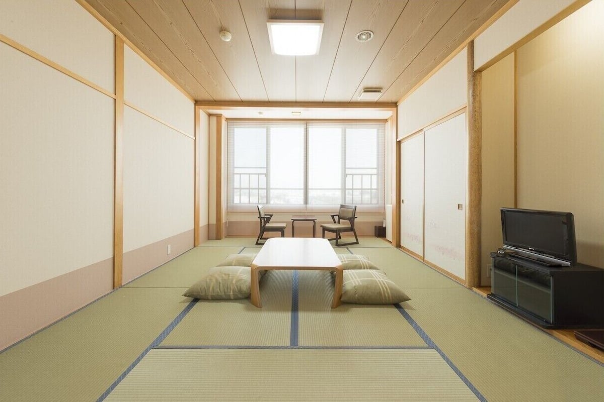 A room with tatami flooring, a low table, chairs, a television, and a large window.