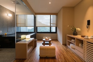 A modern hotel room with a large window, a kitchen area, a small dining table, and a seating area with a wicker ottoman. - Asia Pacific Hotel Beitou (Taipei)