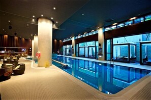 A modern indoor swimming pool with clear blue water, surrounded by lounge areas with seating and a view of the outdoors through large windows. - Asia Pacific Hotel Beitou (Taipei)