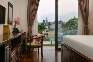 Habitación de hotel con una cama, un escritorio con una computadora portátil, una silla y vistas a edificios y montañas a través de amplios ventanales.