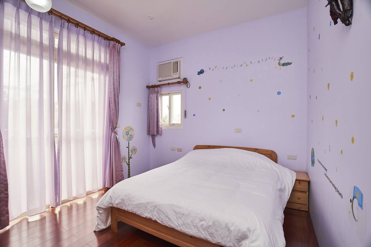 A bedroom with a wooden bed, a window with purple curtains, an air conditioning unit, and wall decorations.