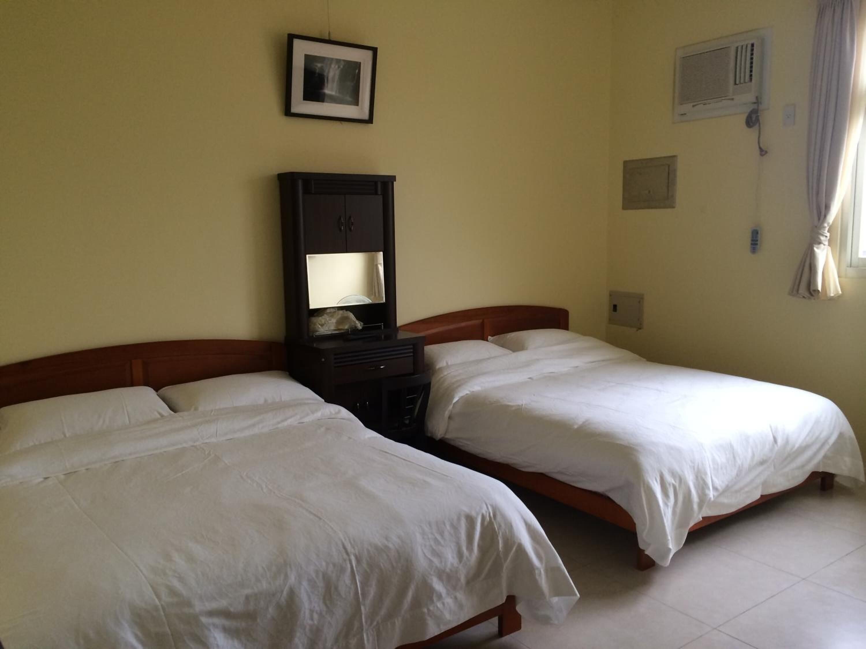 A hotel room with two single beds, a wooden headboard, a nightstand with a mirror, and a wall-mounted air conditioning unit.