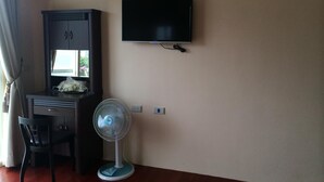 Una habitación con un escritorio, una silla, un televisor montado en la pared y un ventilador de pie.