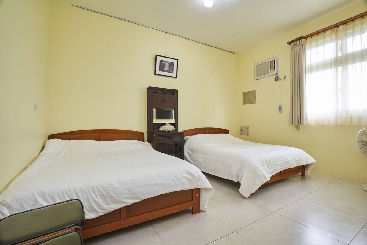 A room with two single beds, a desk with a chair, a picture on the wall, a window with curtains, and a wall-mounted fan.