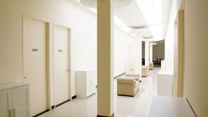 Un corredor moderno de hotel con paredes, puertas y mobiliario blancos. Se observan señales con números de habitación y una recepción al fondo.