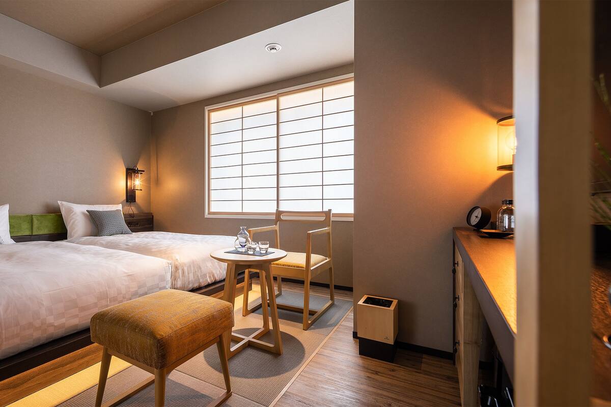 A hotel room with two beds, a small table with chairs, a window with blinds, and a plant on the right side.