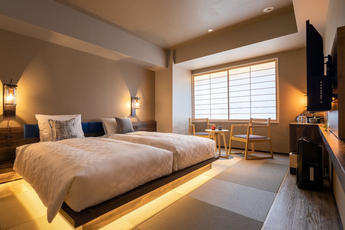 A hotel room with a large bed, a desk, a chair, a TV, and a window with blinds.