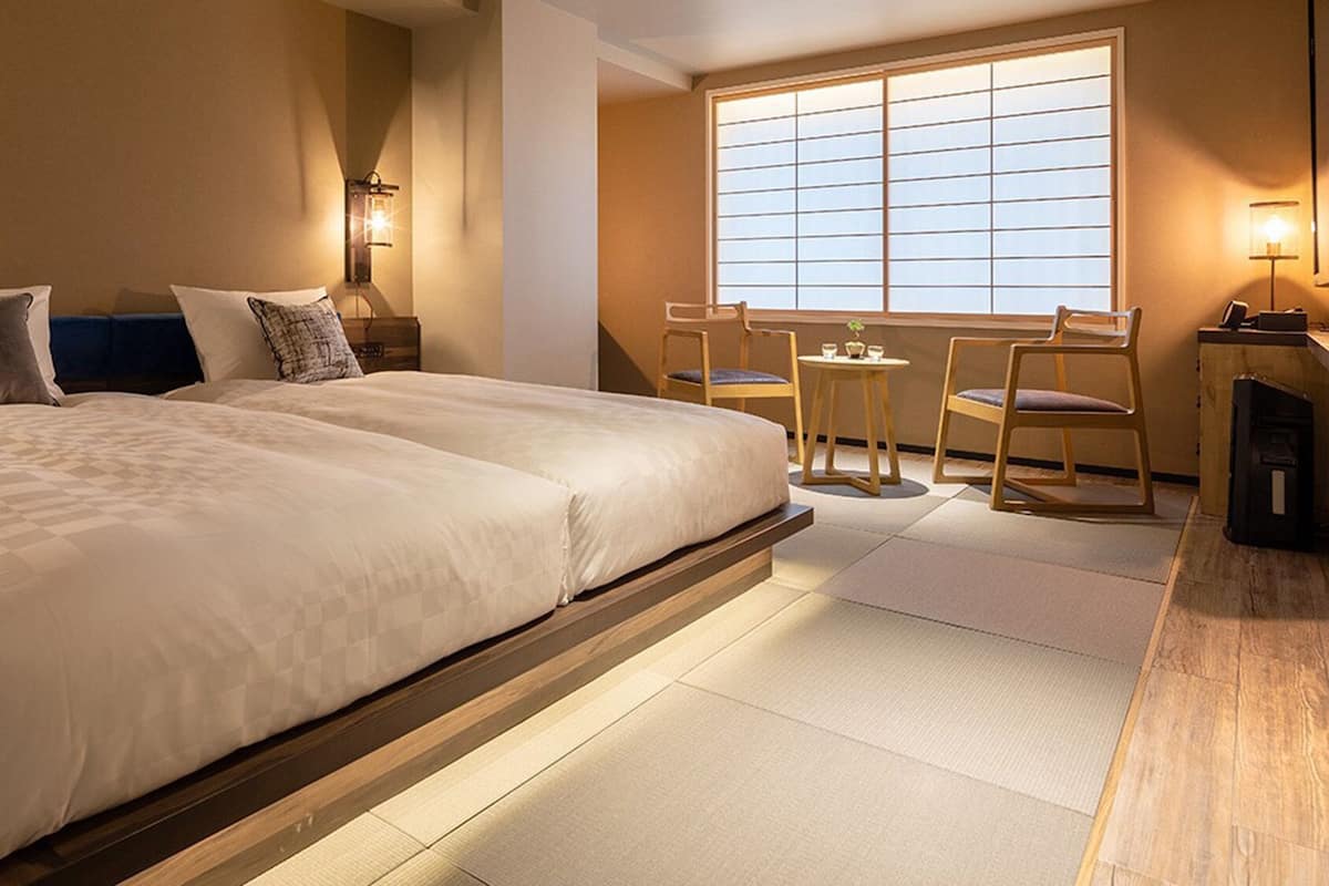 Quarto de hotel com uma cama grande, uma mesinha com cadeiras e vista pela janela.