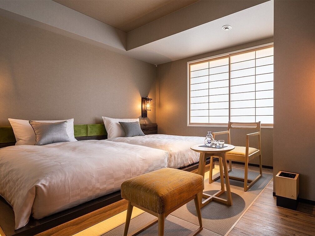 Quarto de hotel com duas camas, uma mesa pequena com cadeiras, uma janela com persianas e uma lixeira.