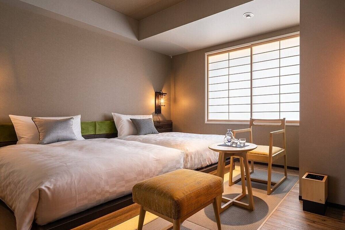 A hotel room with two beds, a small table with chairs, a window with blinds, and a trash bin.