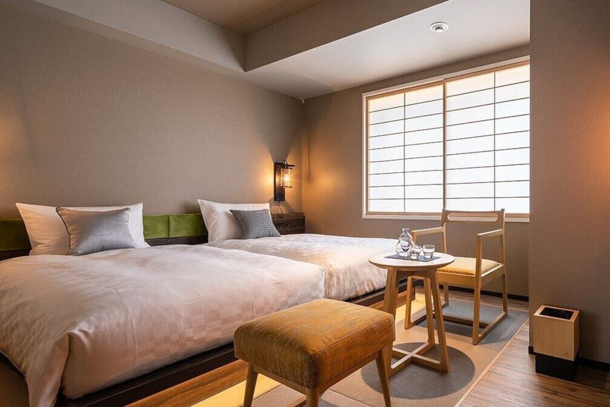 Quarto de hotel com duas camas, uma mesa pequena com cadeiras, uma janela com persianas e uma lixeira.